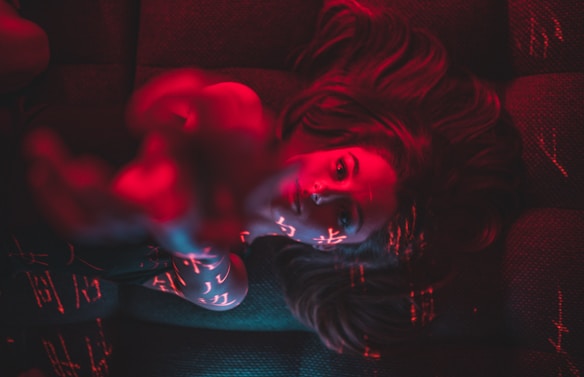 A person with long hair is lying down, with red and blue lighting casting intricate patterns and symbols across their face and body. The lighting creates a mysterious and artistic atmosphere.
