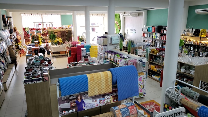 A well-organized store interior with neatly arranged products. Various home goods and decorations are displayed, including kitchenware, artificial flowers, and textiles. Shelves are filled with neatly stacked items, and the store is brightly lit with natural and artificial light.