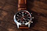 A sleek chronograph watch with a detailed dial featuring three sub-dials and a mix of red and white details. The face has a metallic border with engraved numbers and a brown leather strap, all set against a textured wooden surface.