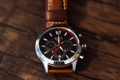 A sleek chronograph watch with a detailed dial featuring three sub-dials and a mix of red and white details. The face has a metallic border with engraved numbers and a brown leather strap, all set against a textured wooden surface.