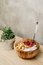 A wooden bowl containing a mixture of cashews, yogurt, and berries, placed on a light wooden surface. A spoon rests inside the bowl, and beside it is a potted plant with green foliage in a ceramic pot.