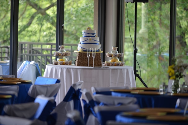 A tiered cake with blue ribbon decoration is displayed on a table covered with a white tablecloth. Surrounding the cake are neatly arranged cupcakes in decorative wrappers. The setting appears to be an elegant indoor event space with large windows offering a view of lush greenery outside. Chairs in the foreground are adorned with blue and white fabric covers, contributing to a festive atmosphere.