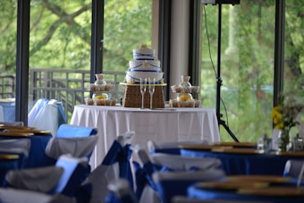 A tiered cake with blue ribbon decoration is displayed on a table covered with a white tablecloth. Surrounding the cake are neatly arranged cupcakes in decorative wrappers. The setting appears to be an elegant indoor event space with large windows offering a view of lush greenery outside. Chairs in the foreground are adorned with blue and white fabric covers, contributing to a festive atmosphere.