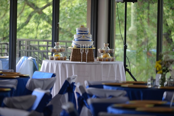 A tiered cake with blue ribbon decoration is displayed on a table covered with a white tablecloth. Surrounding the cake are neatly arranged cupcakes in decorative wrappers. The setting appears to be an elegant indoor event space with large windows offering a view of lush greenery outside. Chairs in the foreground are adorned with blue and white fabric covers, contributing to a festive atmosphere.