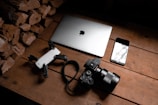 A wooden surface is arranged with a stack of firewood on one side, a laptop with an Apple logo, a smartphone with a reflective screen, a camera with a large lens, and a small drone. The objects are neatly placed, suggesting a theme of technology and photography.