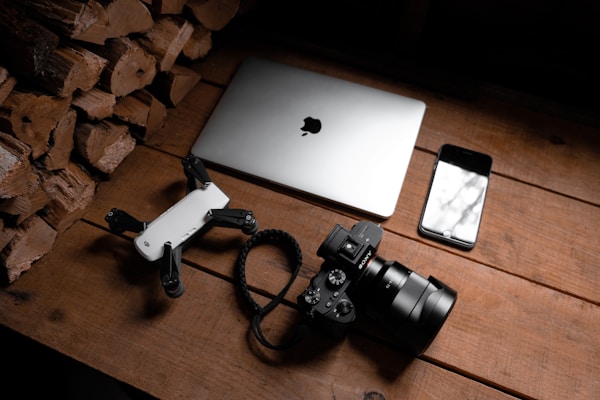 A wooden surface is arranged with a stack of firewood on one side, a laptop with an Apple logo, a smartphone with a reflective screen, a camera with a large lens, and a small drone. The objects are neatly placed, suggesting a theme of technology and photography.