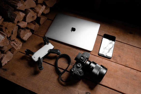 A wooden surface is arranged with a stack of firewood on one side, a laptop with an Apple logo, a smartphone with a reflective screen, a camera with a large lens, and a small drone. The objects are neatly placed, suggesting a theme of technology and photography.