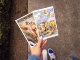 A hand with light blue nail polish holds two art postcards featuring classical paintings. The left postcard displays a scene with mythological figures and a lush background, while the right postcard shows figures and cherubs in a dynamic sky and sea scene. The background includes part of a pathway and grass.