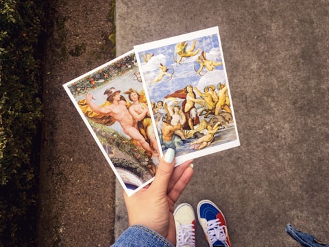 A hand with light blue nail polish holds two art postcards featuring classical paintings. The left postcard displays a scene with mythological figures and a lush background, while the right postcard shows figures and cherubs in a dynamic sky and sea scene. The background includes part of a pathway and grass.