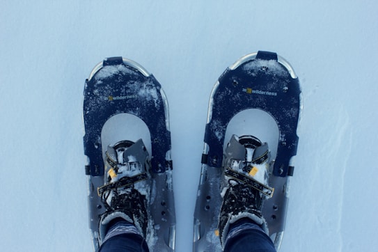 A close-up view of two snowshoes on a snow-covered ground, with a pair of boots strapped into them. The snowshoes are dark-colored with visible snow accumulated on them. The word 'wilderness' is printed on each snowshoe.