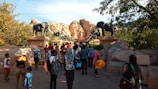A group of people are walking towards an entrance adorned with large elephant statues. The setting is outdoors with rocky hills and trees providing a natural backdrop. The people are dressed casually, suggesting a leisurely outing, and some are carrying backpacks and colorful floatation devices.