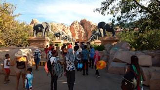 A group of people are walking towards an entrance adorned with large elephant statues. The setting is outdoors with rocky hills and trees providing a natural backdrop. The people are dressed casually, suggesting a leisurely outing, and some are carrying backpacks and colorful floatation devices.
