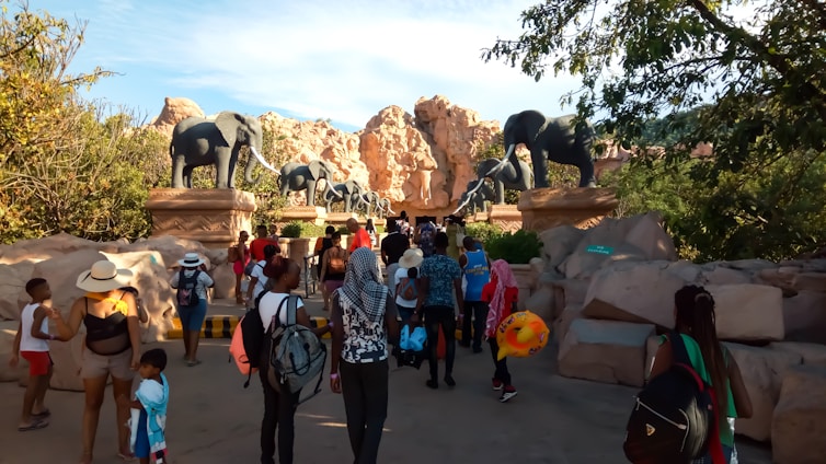 A group of people are walking towards an entrance adorned with large elephant statues. The setting is outdoors with rocky hills and trees providing a natural backdrop. The people are dressed casually, suggesting a leisurely outing, and some are carrying backpacks and colorful floatation devices.