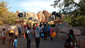 A group of people are walking towards an entrance adorned with large elephant statues. The setting is outdoors with rocky hills and trees providing a natural backdrop. The people are dressed casually, suggesting a leisurely outing, and some are carrying backpacks and colorful floatation devices.