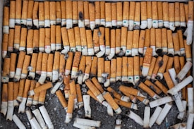 A large collection of cigarette butts arranged neatly in rows on a dark surface. The butts are mostly orange with white filter ends, showing various brands and some charred marks.