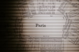 A vintage train schedule centered around the name Paris, with various station names and times laid out in a radial pattern. The text is printed in a dark, classic font on a beige, slightly translucent background that has subtle horizontal lines.