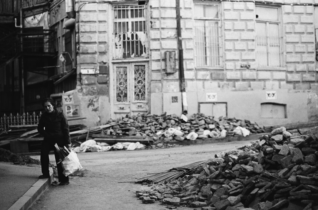 grayscale photo of man in black jacket and pants sitting on concrete floor