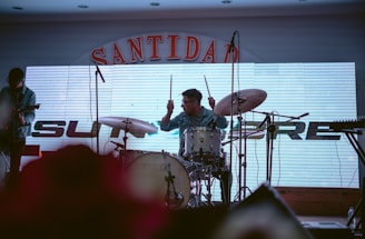 A drummer is energetically playing on a drum set with a cymbal while a guitarist stands nearby. The backdrop features a large electronic display with part of the word 'SANTIDAD' visible on top and some additional lettering partially obscured. Microphones are set up in front of the musicians, and the lighting creates a dim atmosphere.
