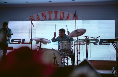 A drummer is energetically playing on a drum set with a cymbal while a guitarist stands nearby. The backdrop features a large electronic display with part of the word 'SANTIDAD' visible on top and some additional lettering partially obscured. Microphones are set up in front of the musicians, and the lighting creates a dim atmosphere.