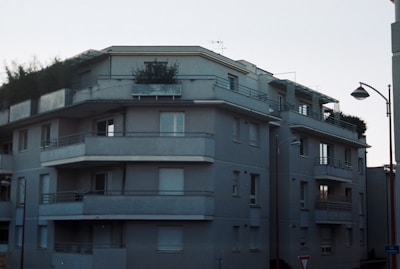 A multi-story residential building with a modern architectural design. The structure features several balconies, each with metal railing, and a flat roof with greenery visible. The facade is painted in a uniform gray tone, and a street light is positioned at the corner. Windows vary between open and covered with shutters.