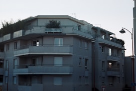 A multi-story residential building with a modern architectural design. The structure features several balconies, each with metal railing, and a flat roof with greenery visible. The facade is painted in a uniform gray tone, and a street light is positioned at the corner. Windows vary between open and covered with shutters.