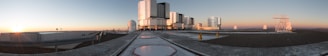 A panoramic view of an observatory with several large telescope domes and rectangular structures situated on a flat rooftop. The sun is setting on the horizon, casting a warm glow across the scene, and the sky transitions from blue to orange. The foreground features smooth, light-colored pathways and darker areas suggesting gravel or similar material.