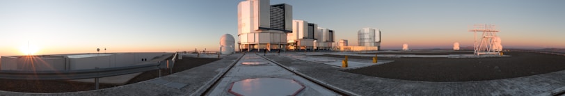 A panoramic view of an observatory with several large telescope domes and rectangular structures situated on a flat rooftop. The sun is setting on the horizon, casting a warm glow across the scene, and the sky transitions from blue to orange. The foreground features smooth, light-colored pathways and darker areas suggesting gravel or similar material.