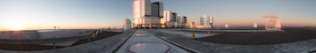 A panoramic view of an observatory with several large telescope domes and rectangular structures situated on a flat rooftop. The sun is setting on the horizon, casting a warm glow across the scene, and the sky transitions from blue to orange. The foreground features smooth, light-colored pathways and darker areas suggesting gravel or similar material.