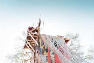 A handcrafted, elevated wooden structure with a colorful facade is set against a clear sky. The structure incorporates art and writing on its surface, with netting draped over one side. Sparse trees can be seen in the background, emphasizing an open, airy environment.
