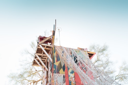 A handcrafted, elevated wooden structure with a colorful facade is set against a clear sky. The structure incorporates art and writing on its surface, with netting draped over one side. Sparse trees can be seen in the background, emphasizing an open, airy environment.