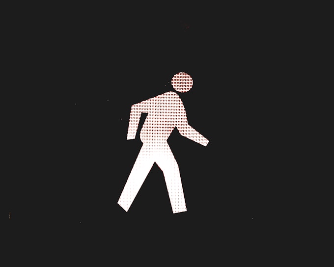 A white stick figure walking symbol is depicted against a black background. The stick figure is composed of a pattern of small repeating characters that resemble the letter R. The figure is shown in mid-step with one leg forward and one arm bent at the elbow.