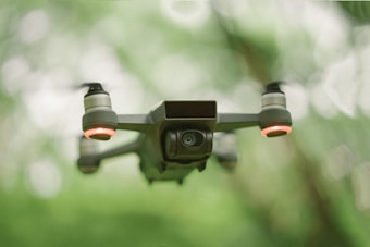 A drone with red-lit propellers is captured in flight against a blurred green and white background, giving an impression of being outdoors, possibly in a forest or garden.