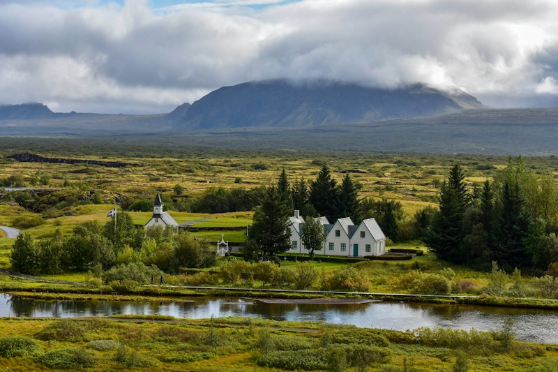 White-gabled farmhouses and a small white church beside a river in a green rift valley, with a cloud-capped mountain behind