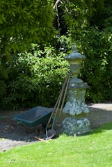 A garden scene featuring a weathered stone pedestal with an ornate design. Leaning against it are three gardening tools, including a shovel and a rake. Next to the pedestal is a green wheelbarrow, partially filled with soil. Behind the setup, lush green foliage creates a dense backdrop, casting a mix of light and shadow on the ground.