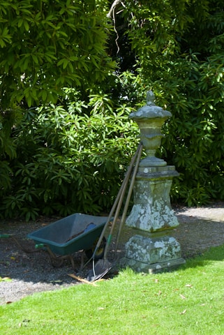 A garden scene featuring a weathered stone pedestal with an ornate design. Leaning against it are three gardening tools, including a shovel and a rake. Next to the pedestal is a green wheelbarrow, partially filled with soil. Behind the setup, lush green foliage creates a dense backdrop, casting a mix of light and shadow on the ground.