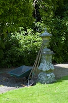 A garden scene featuring a weathered stone pedestal with an ornate design. Leaning against it are three gardening tools, including a shovel and a rake. Next to the pedestal is a green wheelbarrow, partially filled with soil. Behind the setup, lush green foliage creates a dense backdrop, casting a mix of light and shadow on the ground.