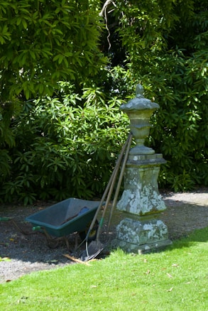 A garden scene featuring a weathered stone pedestal with an ornate design. Leaning against it are three gardening tools, including a shovel and a rake. Next to the pedestal is a green wheelbarrow, partially filled with soil. Behind the setup, lush green foliage creates a dense backdrop, casting a mix of light and shadow on the ground.