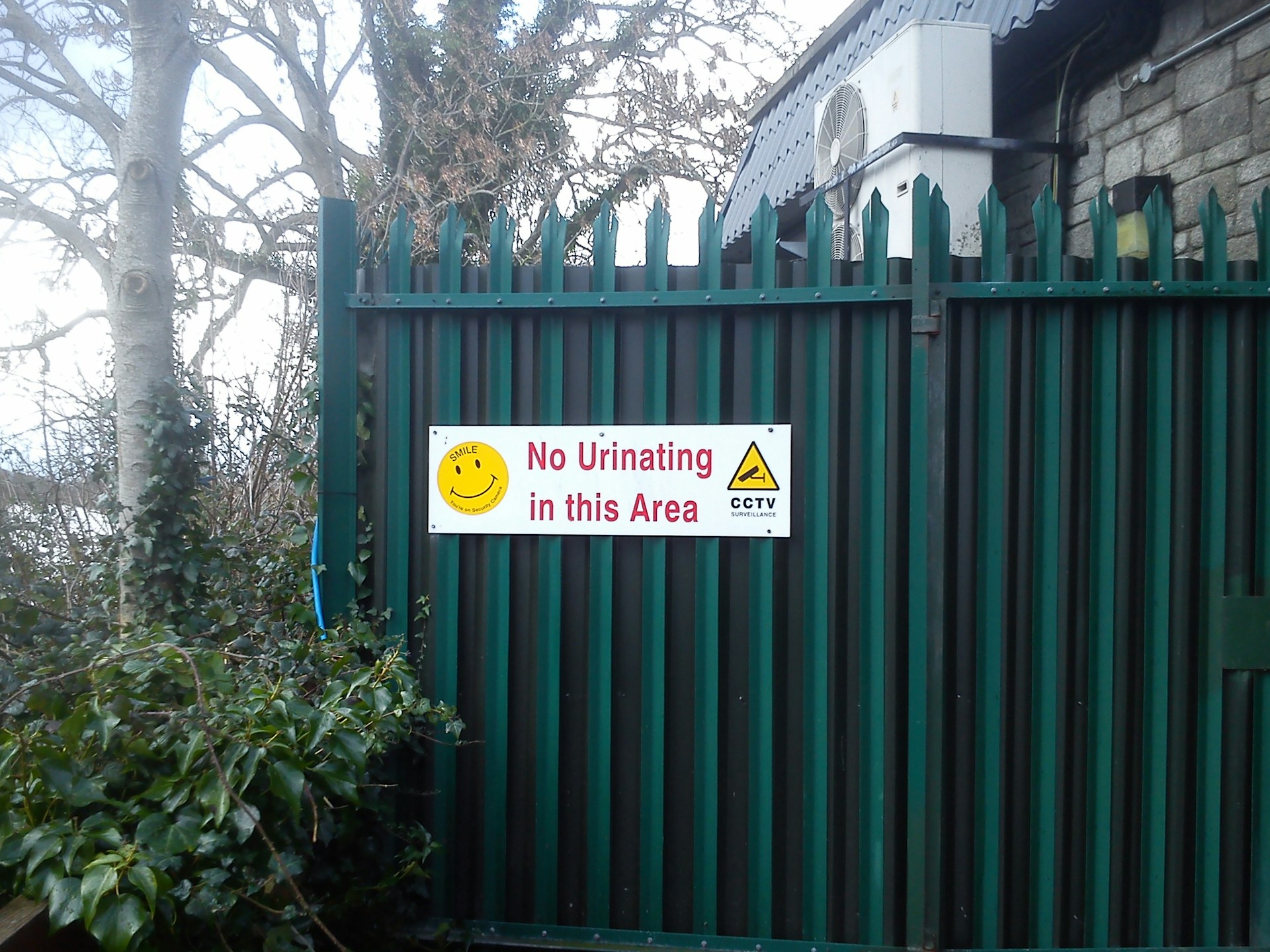 A metal fence with vertical bars is shown, in front of a building with a stone wall and an air conditioning unit. A sign attached to the fence reads 'No Urinating in this Area' alongside a yellow smiley face and warning symbols indicating CCTV surveillance. Lush greenery and trees are visible around the fence.