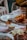 A dining table is set with various food items, including a plate of sliced cheese and cured meats, a jar containing a white substance tied with a ribbon, and dishes topped with slices of fruit. A wine glass is positioned prominently on the table against a pastel tablecloth.