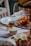 A dining table is set with various food items, including a plate of sliced cheese and cured meats, a jar containing a white substance tied with a ribbon, and dishes topped with slices of fruit. A wine glass is positioned prominently on the table against a pastel tablecloth.