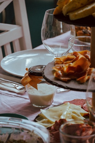 A dining table is set with various food items, including a plate of sliced cheese and cured meats, a jar containing a white substance tied with a ribbon, and dishes topped with slices of fruit. A wine glass is positioned prominently on the table against a pastel tablecloth.