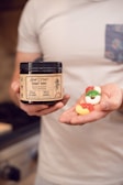 A person holds a jar labeled 'Hemp Extract Gummy Rings' in one hand and several colorful gummy rings in the other. The jar features details such as being multi-flavored and full-spectrum.
