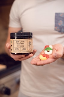 A person holds a jar labeled 'Hemp Extract Gummy Rings' in one hand and several colorful gummy rings in the other. The jar features details such as being multi-flavored and full-spectrum.