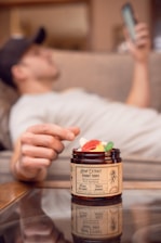 A person is relaxing on a couch with a phone in hand, while a jar of hemp extract gummy rings is in the foreground. The focus is on the jar, with colorful gummies visible inside.
