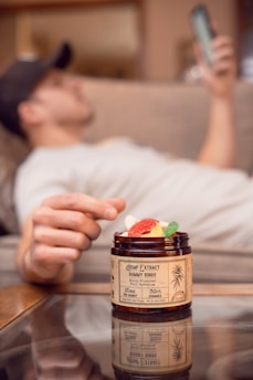 A person is relaxing on a couch with a phone in hand, while a jar of hemp extract gummy rings is in the foreground. The focus is on the jar, with colorful gummies visible inside.