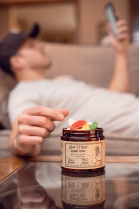 A person is relaxing on a couch with a phone in hand, while a jar of hemp extract gummy rings is in the foreground. The focus is on the jar, with colorful gummies visible inside.