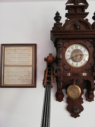 A vintage wall clock with ornate wooden carvings and a pendulum next to a framed sheet of music. The clock features Roman numerals on its face and intricate decorative elements. It is alongside the neck and strings of a stringed instrument, likely a cello or double bass, leaning against the clock.