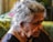 An older woman is sitting in profile, with grey curly hair and a nose stud. She is wearing a dark-colored garment and has a gold necklace. The lighting is soft, with a blurred background that is mostly green and yellow.