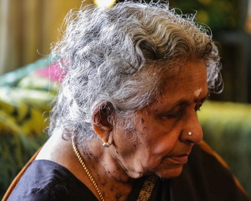 An older woman is sitting in profile, with grey curly hair and a nose stud. She is wearing a dark-colored garment and has a gold necklace. The lighting is soft, with a blurred background that is mostly green and yellow.