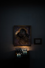 A religious icon of the Virgin Mary holding baby Jesus is mounted on a dimly lit wall. Beneath the icon, several candles are lit, casting a soft, warm glow. There is a small framed plaque on the wall beside the icon.